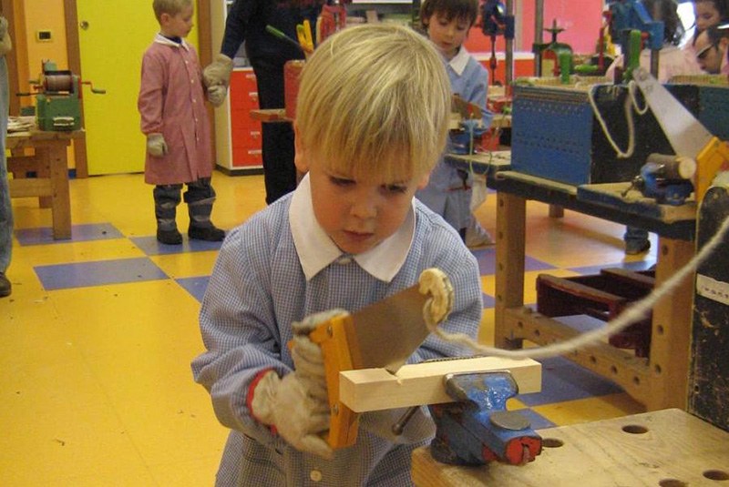 bambino biondo col grambiule che usa la sega per tagliare un legno